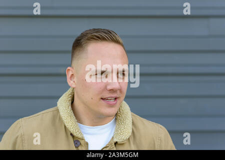 Thoughtful attractive young man looking away to the side with a quiet smile outdoors against a grey wall Stock Photo