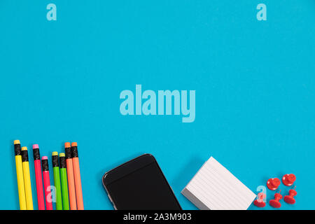 Multicolored pencils, black mobile phone, a pile of white scratch paper and a group of red pushpins are half visible lying on a pale-blue surface Stock Photo