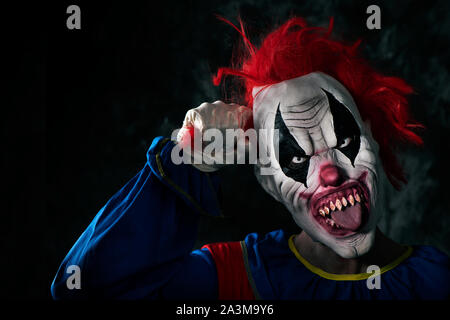 closeup of a scary evil clown with white eyes and bloody teeth, sticking out his tongue while is grabbing his red hair and staring at the observer wit Stock Photo