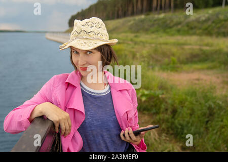 Young beautiful smiling woman is holding smart phone in hand and looking to the camera. Forest and lake on a background. Stock Photo