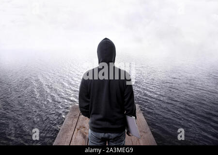 back view of hacker in the hood, with laptop in the hand, on wooden pier going into the water and fog, programming difficulties concept Stock Photo