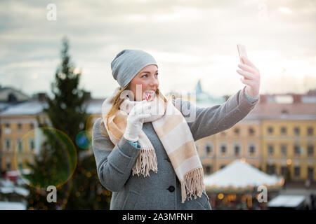 Young woman in Christmas market eating candy cane and taking selfie. Girl in knitted warm hat and scarf. Illuminated and decorated fair kiosks, shops Stock Photo