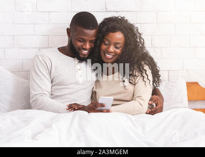 Smiling black couple using smartphone sitting on bed together Stock Photo
