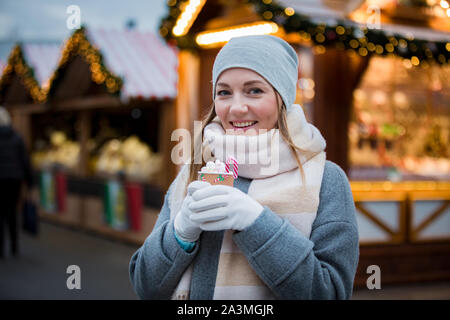 Young woman in Christmas market drinking cup of hot chocolate with marshmallow wearing knitted warm hat and scarf. Illuminated and decorated shops Stock Photo