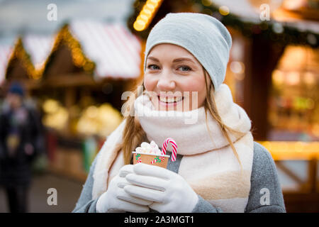 Young woman in Christmas market drinking cup of hot chocolate with marshmallow wearing knitted warm hat and scarf. Illuminated and decorated shops Stock Photo