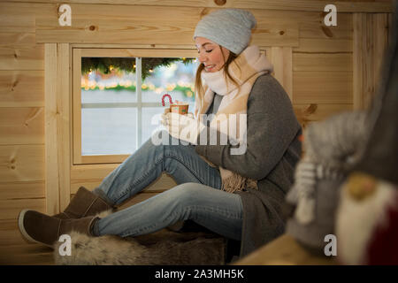Young woman drinking cup of hot chocolate with marshmallow wearing knitted warm hat and scarf. Sitting next to window in Finnish wooden hut Stock Photo