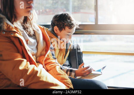 Children students in yellow jackets riding in the city bus. Boy reading a book at the window Stock Photo