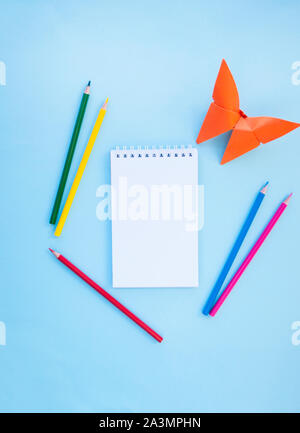 Colorful pencils, a notebook and a paper butterfly on blue background with copyspace. Flat lay style. Back to school concept. Stock Photo