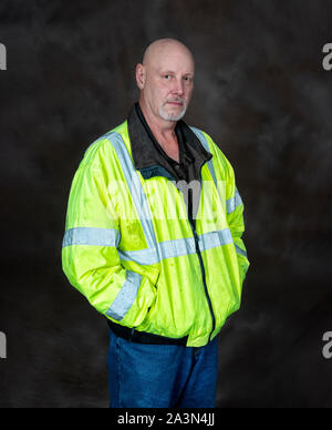 Vertical shot of a mature male worker in a reflective coat on a brown background.  He is standing with his hands in his pockets. Stock Photo