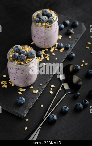 Healthy breakfast concept. Jars of homemade yogurt with blueberries and oats on black background. Vertical. Stock Photo