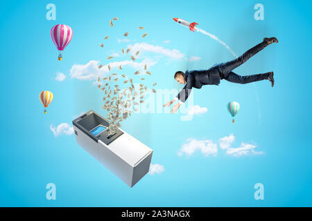 Businessman reaching to ATM machine with dollars flying out, hot air balloons and silver red space rocket in the air on blue background Stock Photo