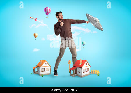 Giant young man with two small houses at his feet, standing in half-turn and fighting a UFO. Stock Photo