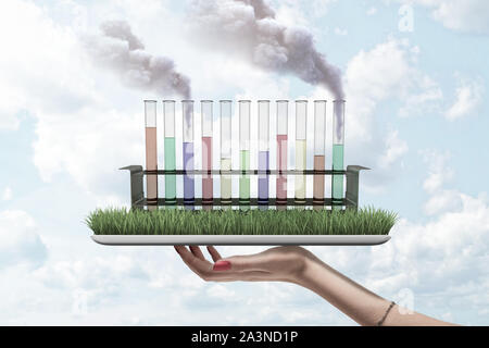 Test tubes filled with differently colored liquids, two tubes spouting smoke, standing on grass-covered screen of digital tablet held in woman's hand. Stock Photo