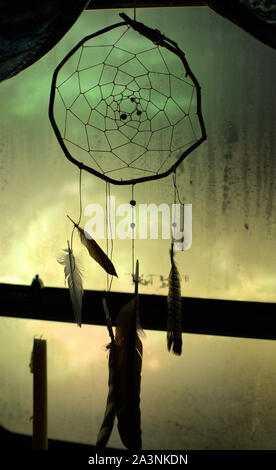 A simple handcrafted dreamcatcher decorating a window, with dark colors contrasting with the green and yellow blurry background Stock Photo