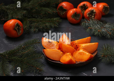 Slices of ripe orange persimmon are on a wooden plate on a dark background, horizontal orientation, closeup