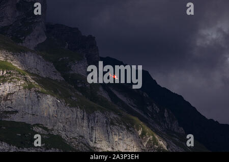 Red helicopter on the background of the massive dark mountain. Overcast sky. Rescue helicopter in mountains, dusk. Swiss alpine Mountains. Switzerland. Stock Photo