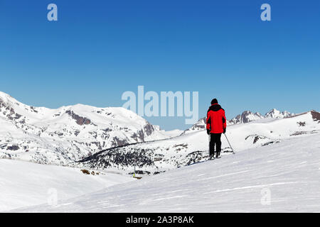 PYRENEES, ANDORRA - FEBRUARY 14, 2019: A skier in a bright red jacket stands on top of a mountain on a winter sunny day. Mountain landscape and blue s Stock Photo