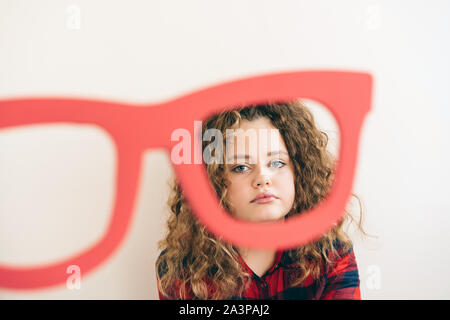 Cute curly long-haired blonde teen girl looks over funny big glasses red color Stock Photo