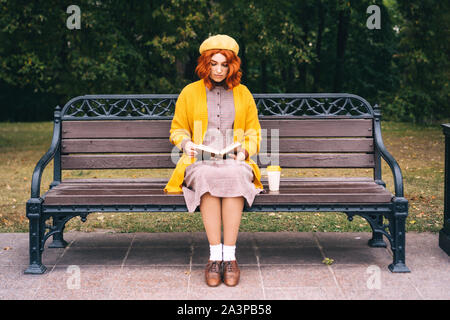 A beautiful red-haired girl with a curly hairstyle is sitting on a park bench Stock Photo