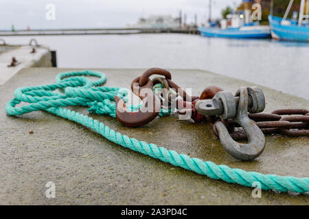 Close up view of rusty hook and green nylon rope on concrete floor of waterside at harbour   in Denmark with blur background of boats and cloudy sky. Stock Photo