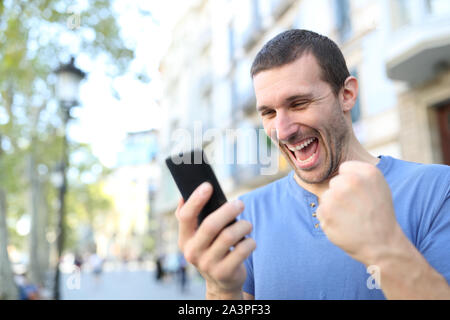 Excited man celebrating good news checking mobile phone content standing in the street Stock Photo