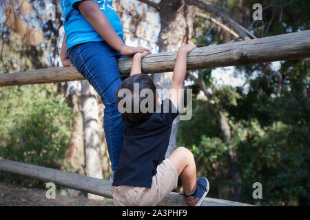 A little brother trying to climb up a wooden fence to sit next to his big sister and enjoy the view in nature. Stock Photo