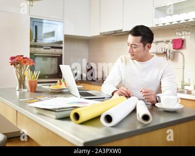 young asian free lance designer sitting at kitchen counter creating a design using pen tablet and laptop computer. Stock Photo