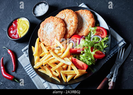delicious lunch, fried turkey burgers served with lettuce tomato salad and french fries on a black plate on a concrete  table, with ketchup and mustar