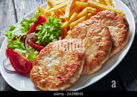 close-up of freshly fried three turkey burgers served with lettuce tomato salad and french fries on a white plate on a rustic wooden table, horizontal Stock Photo