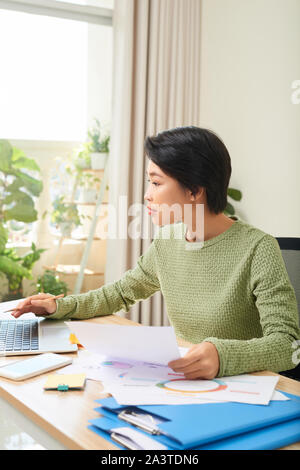 Concentrated Asian woman sit at table working on laptop making notes in notebook at home office Stock Photo