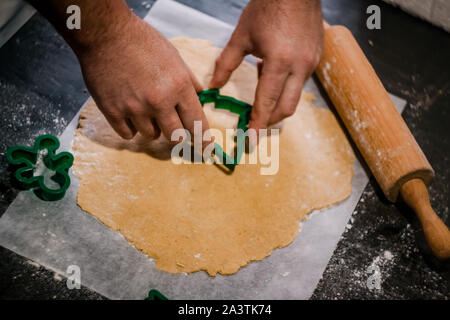 male hands cut out gingerbread cookie in the form of a snowflake, Christmas tree, a man from raw dough on parchment baking paper on a dark background Stock Photo