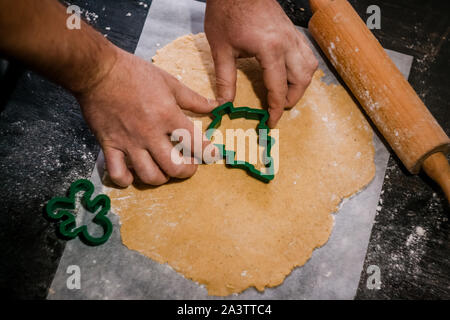 male hands cut out gingerbread cookie in the form of a snowflake, Christmas tree, a man from raw dough on parchment baking paper on a dark background Stock Photo
