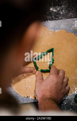male hands cut out gingerbread cookie in the form of a snowflake, Christmas tree, a man from raw dough on parchment baking paper on a dark background Stock Photo