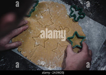 male hands cut out gingerbread cookie in the form of a snowflake, Christmas tree, a man from raw dough on parchment baking paper on a dark background Stock Photo