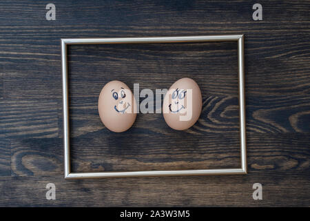 Picture golden frame and two funny eggs smiling on dark wooden wall background, close up. Eggs family emotion face portrait. Couple eggs with happy fa Stock Photo
