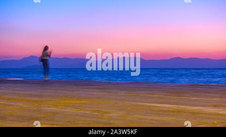 Sunset over Aegean Sea in Aegina Island and a girl on the wharf, Greece, Long exposition, motion blur Stock Photo