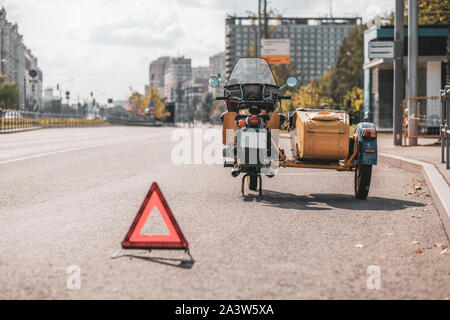 A retro motorcycle with a sidecar without a wheel stands on the edge of the roadway. A warning triangle is placed behind. Sunny autumn day in the city Stock Photo