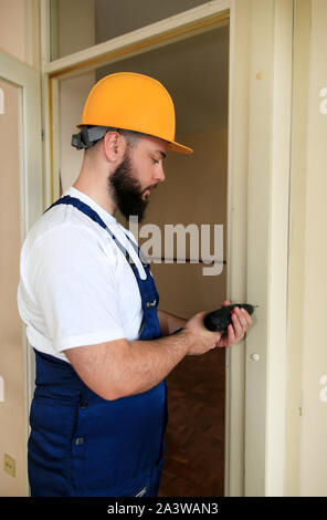 Construction worker and handyman works on renovation of apartment. Builder with electric drill drills nail hole into wooden wall door. Stock Photo