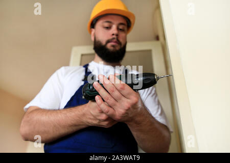 Construction worker and handyman works on renovation of apartment. Builder with electric drill drills nail hole into wooden wall door. Stock Photo