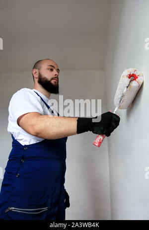 Construction worker doing finish renovation at apartment. Professional painter contractor using white paint roller brush painting of concrete wall. Stock Photo