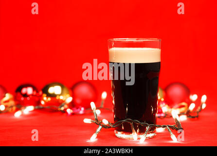 Pint glass of dark beer or stout ale with christmas lights and baubles on red background Stock Photo
