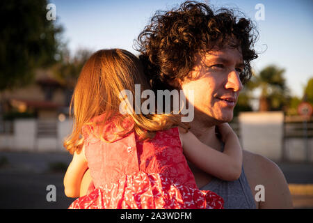 Young father holding his little daughter outdoor at sunset. The man is looking away while the child is back view wearing a red dress. Stock Photo