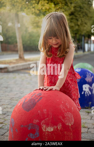 Little child girl playing with red concrete bollard at sunset in urban outdoor location with golden light. Vertical shot. Stock Photo