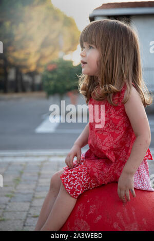 Little child girl sitting on a red concrete bollard at sunset in urban outdoor location with golden light. Vertical shot. Stock Photo