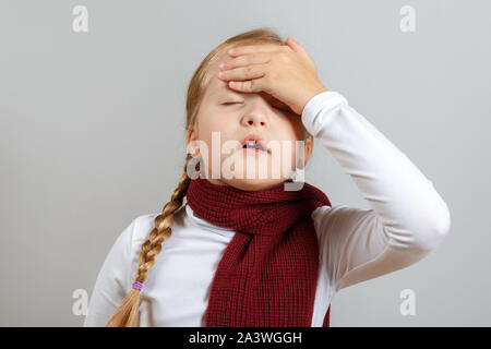 A little girl in a scarf is suffering from an illness. The child holds on to his head. The concept of the season of colds. Stock Photo