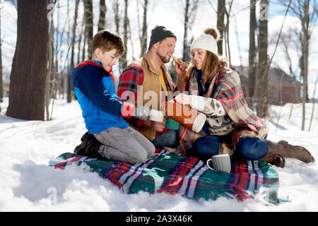 Family of a mother, father and boy child having fun and sharing a cup of cocoa and chocolate chip cookies in snow during the winter season Stock Photo