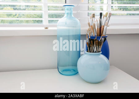 Paint Brushes on a white wooden table, closeup paint Brushes in modern blue vase near the window, modern light interior Stock Photo