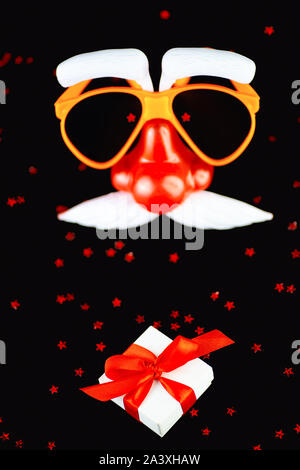 Santa Claus mask and a white gift box with a red ribbon and a scattering of stars on a black background. Stock Photo