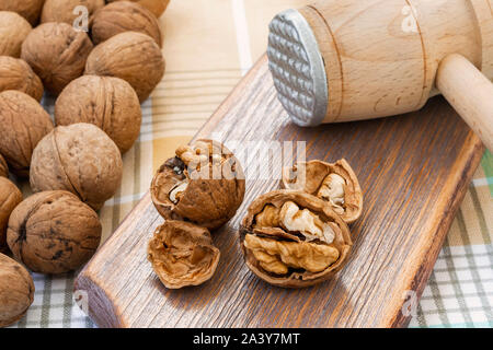 Whole and cracked walnuts (Juglans regia) near wooden meat mallet on a brown wooden board. Natural unbleached nuts. Vegetarian food. Close-up. Stock Photo