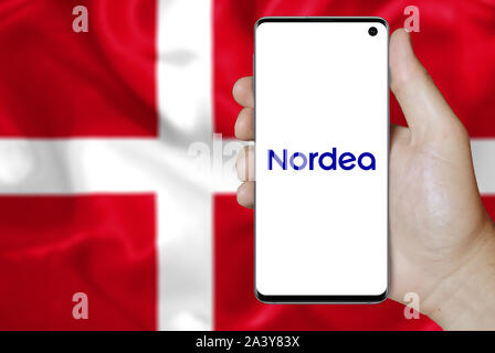 A man holds a phone displaying the logo of company Nordea Bank Abp listed on OMX Copenhagen. Danish flag background. Credit: PIXDUCE Stock Photo